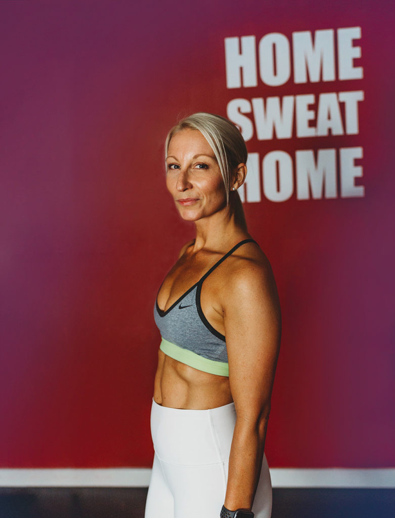 Fitness Instructor Ingrid Segev, wearing fitness apparel, stands in front of "Home Sweat Home" wall sign.